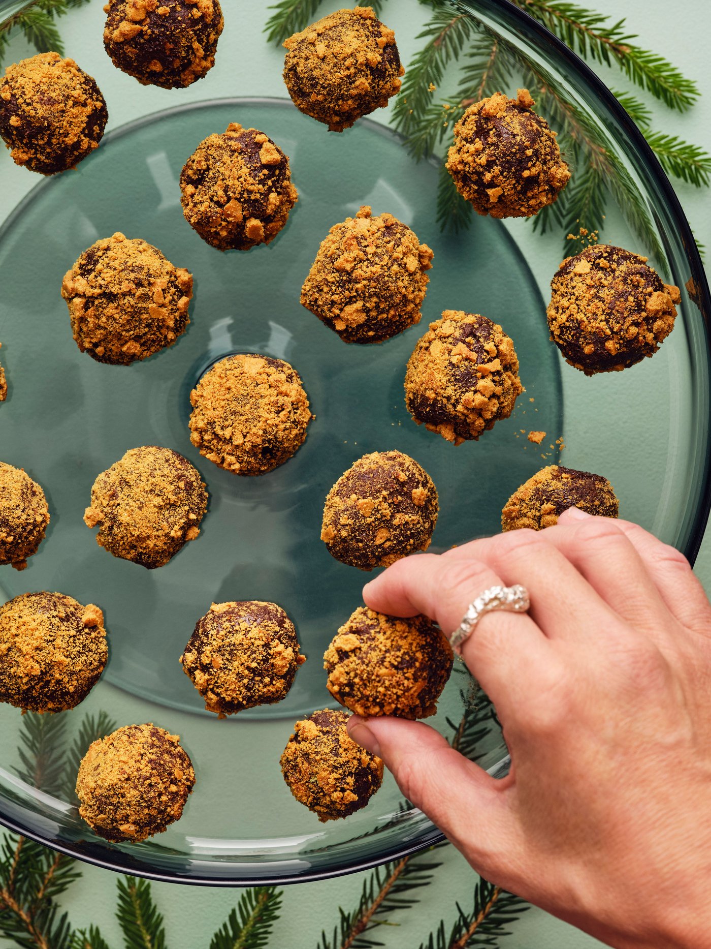 Una mano está cogiendo un dulce de una bandeja de cristal llena de trufas doradas y cubiertas de migas, colocada sobre una superficie verde.