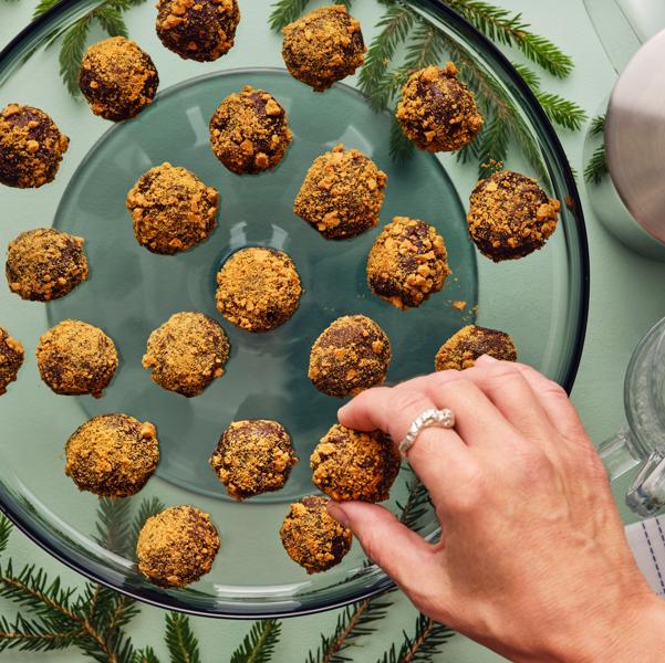 Ei hånd forsyner seg med gyllenbrune sjokoladetrøfler dekket med smuler, fra et glassfat som står på ei grønn overflate.