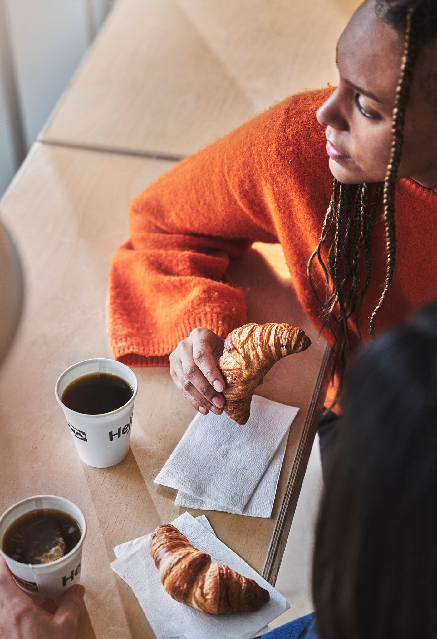 A woman wearing a colour orange sweater is holding a croissant in her hand, and there are two cups of coffee on the table