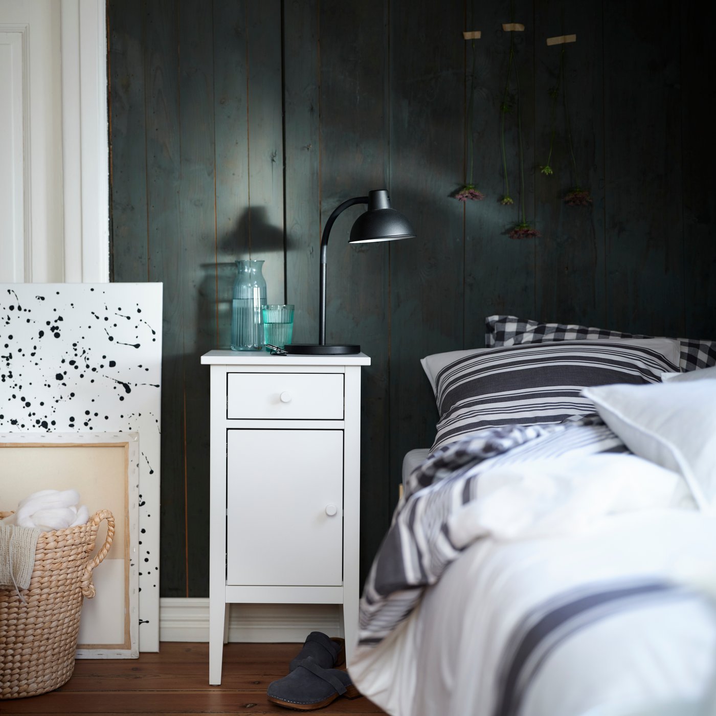 Szafka nocna GULLABERG w kolorze białym ustawiona obok łóżka z pościelą w paski w sypialni z ciemnymi panelami.