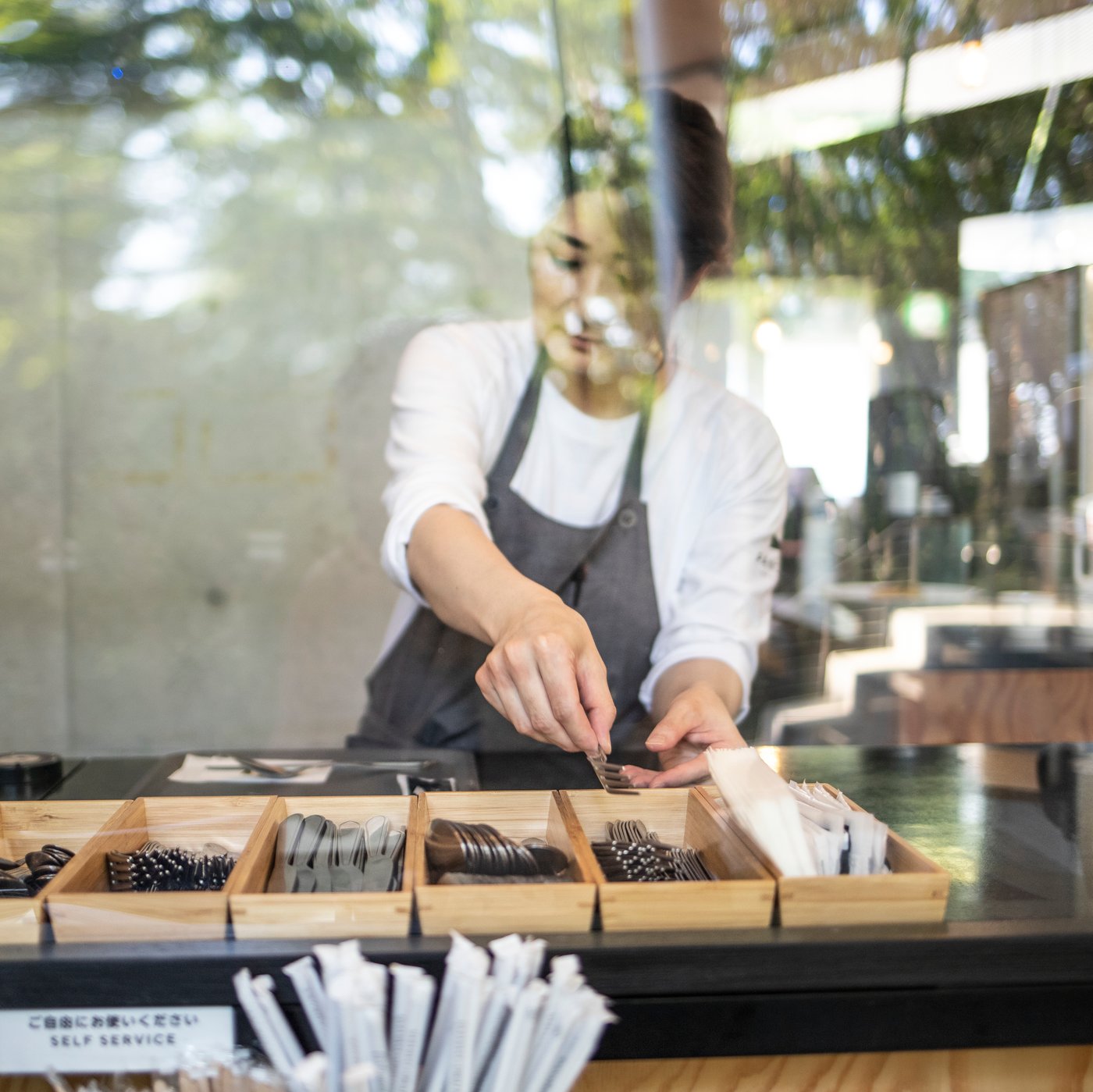 Man schaut durch ein Schaufenster in ein Café. Eine Frau mit einem weißen Langarm Shirt und einer grauen Schürze befüllt gerade Holzboxen mit Besteck.