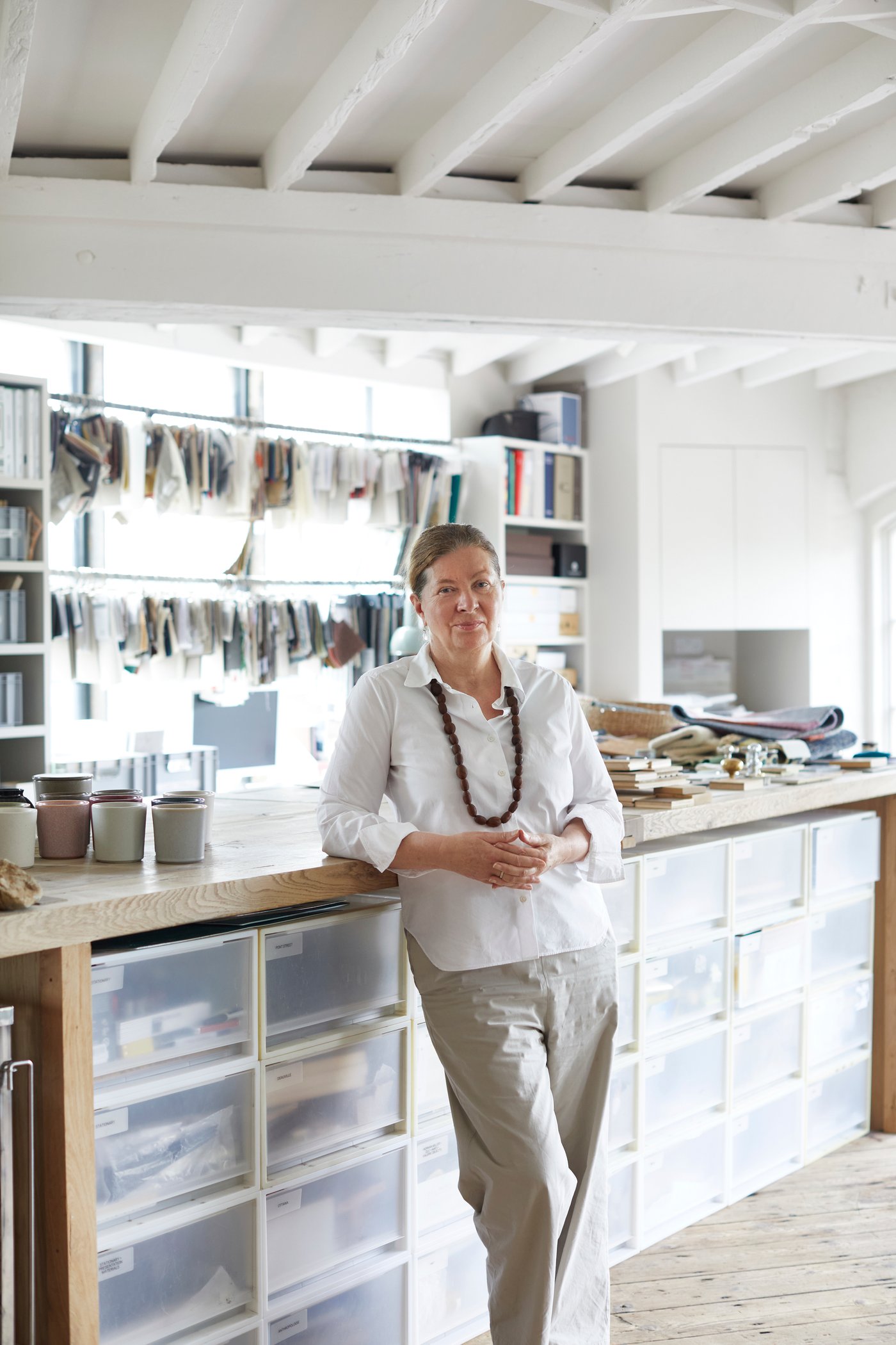 La designer Ilse Crawford, con una camicia bianca e una collana pesante, è appoggiata al piano di lavoro nel suo studio.