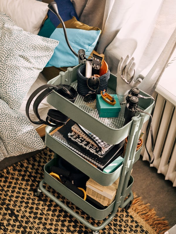 A black, NÄVLINGE LED clamp spotlight is on the top of a grey-green RÅSKOG cart by a bed, with various accessories inside.