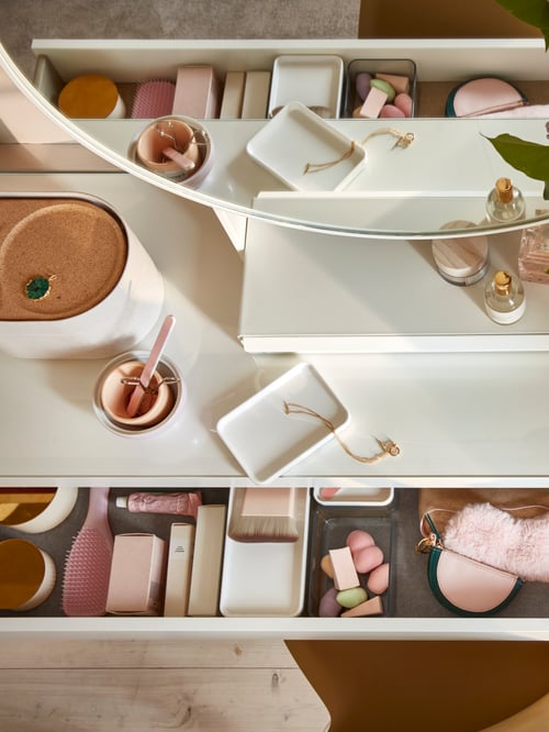 Dressing table seen from above, with a round mirror with white wooden frame on top and a drawer full of makeup products.