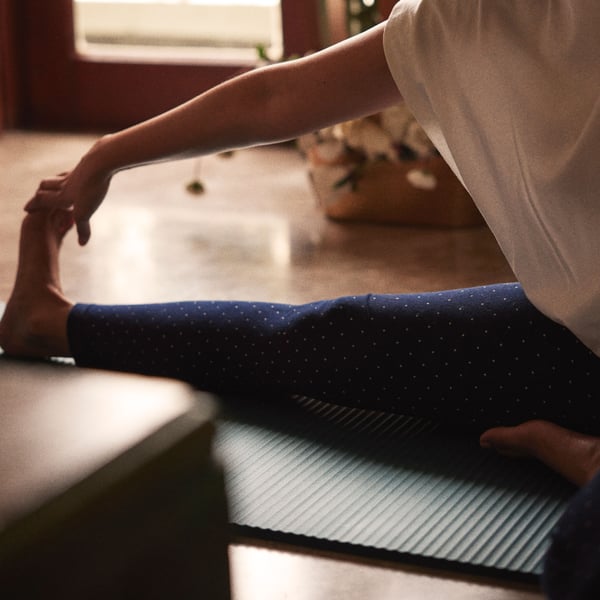A person sits on a yoga mat and stretches out his right leg and reaches his hand towards his foot.