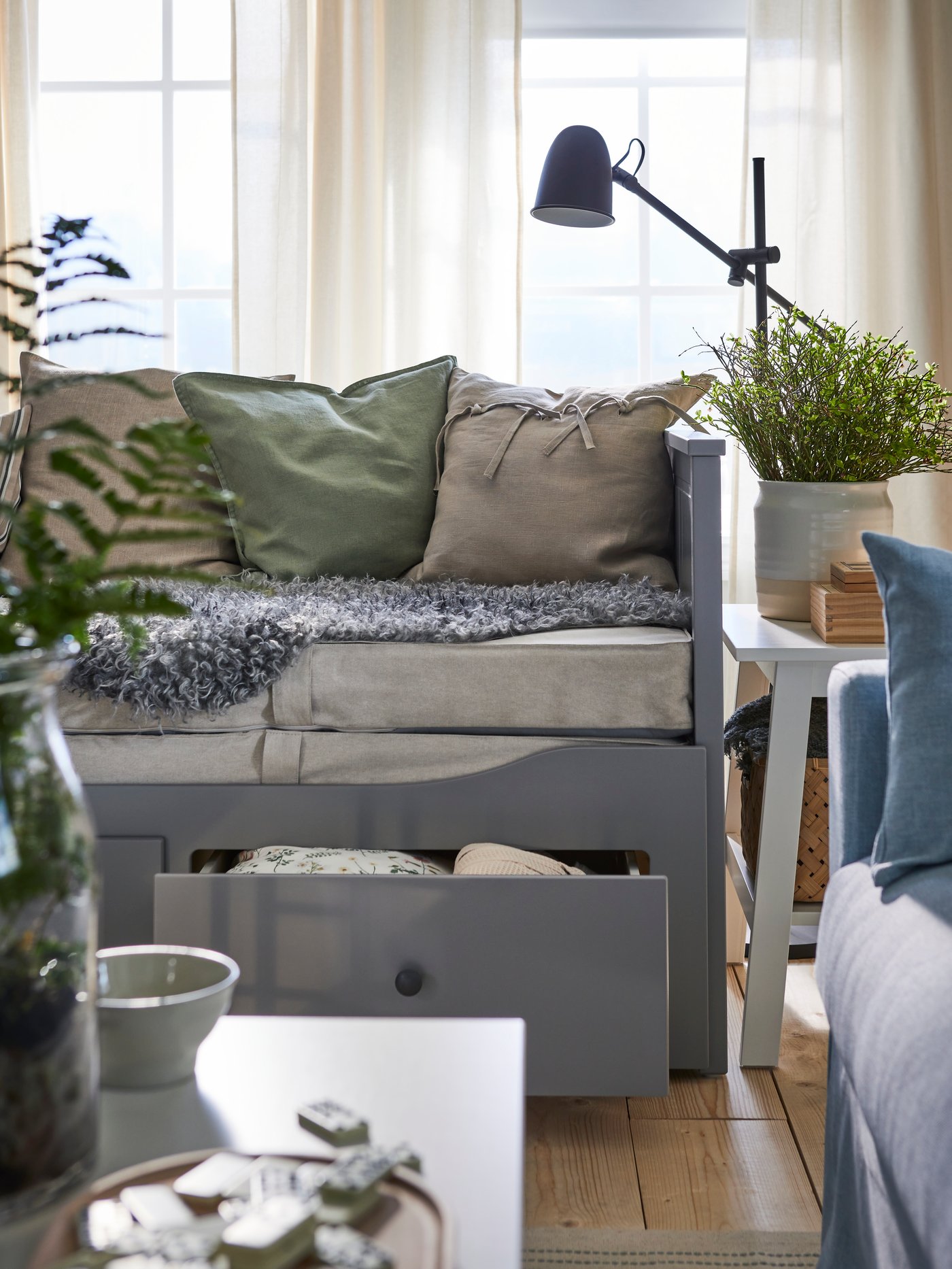 In einem Wohnzimmer im Landhausstil ist ein graues HEMNES Tagesbett mit drei Schubladen zu sehen. Eine Schublade steht offen, auf dem Bett liegen viele Kissen.