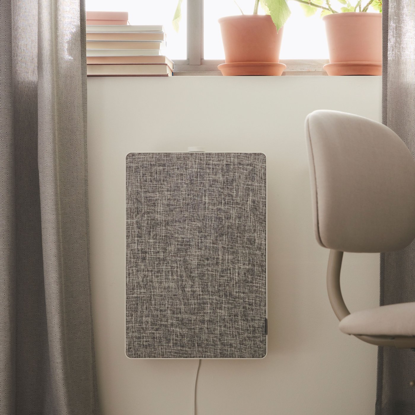 En FÖRNUFTIG luftrenare är monterad på en vägg under ett fönster. På fönsterbrädan står några böcker och växter.