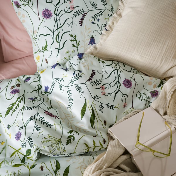 A close-up of a pale blue/floral pattern MATTOXBÄR duvet cover and pillowcase, on a bed with a book, eyeglasses and a throw.