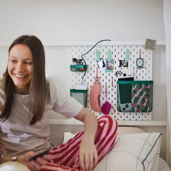 Henkilö istuu sängyllä, jonka päädyssä SKÅDIS-säilytystaulu, jossa on vihreitä kiinnikkeitä ja koreja.