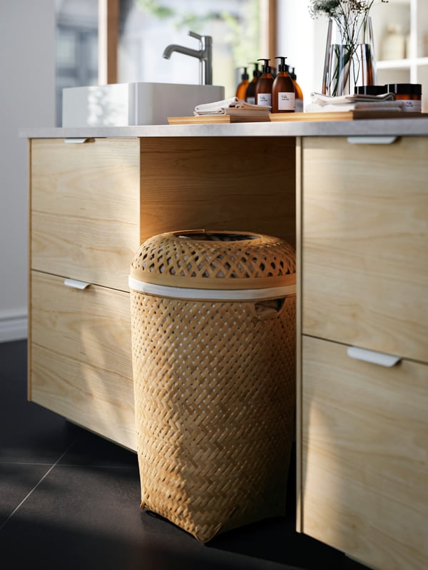 A SALUDING handwoven-bamboo laundry basket placed beneath a worktop and framed by ASKERSUND light-ash-effect drawer fronts.