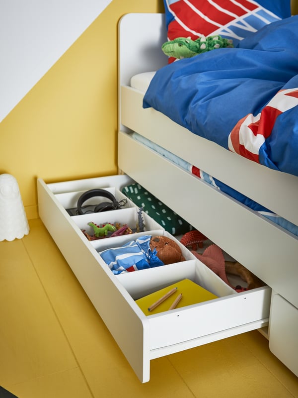A white children's bed with storage underneath revealing colouring materials, toys, bed linen, headphones in tidy boxes.