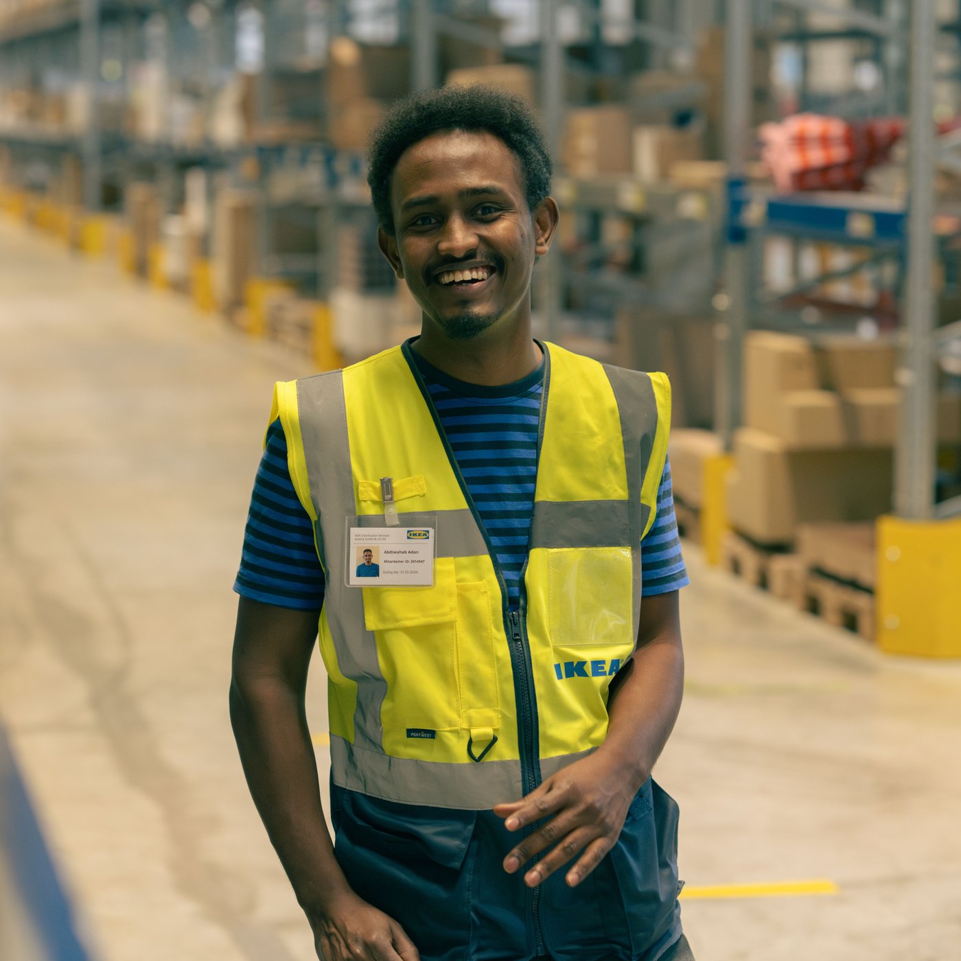 Abdi bei der Arbeit im Warenlager eines IKEA Einrichtungshauses. Er trägt eine gelbe Sicherheitsweste.