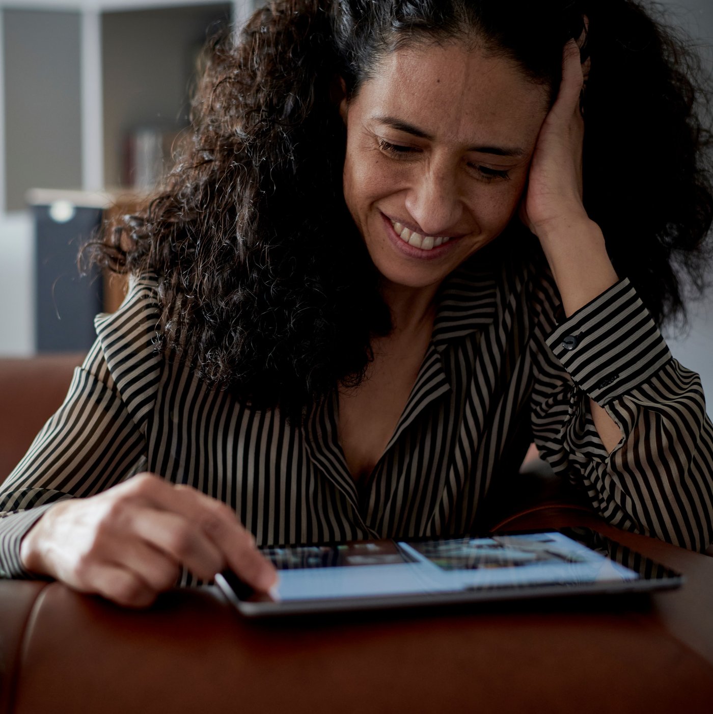 شخص يبتسم يجلس إلى طاولة يسند رأسه على إحدى يديه وينظر في جهاز لوحي على سطح طاولة.