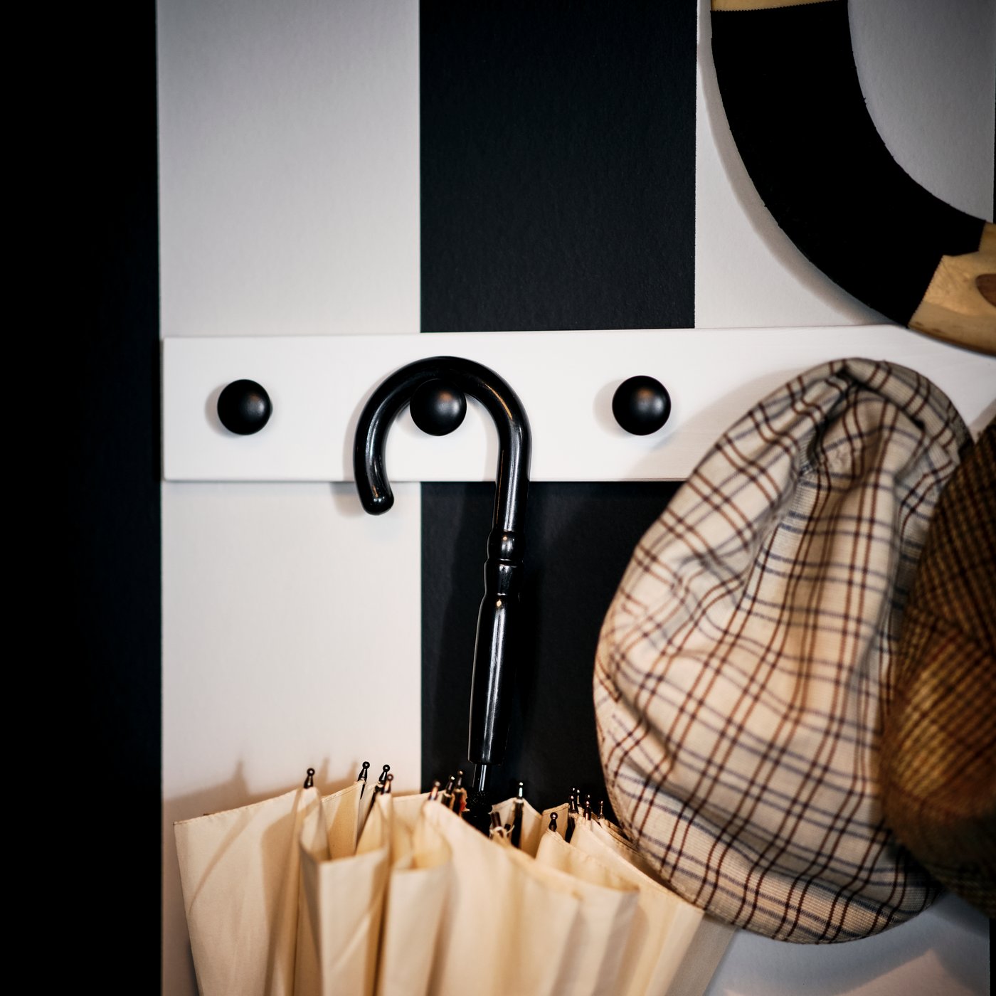 Various outdoor items held by black ENERYDA knobs on a white-stain LURT rack mounted on a black-and-white-striped wall.
