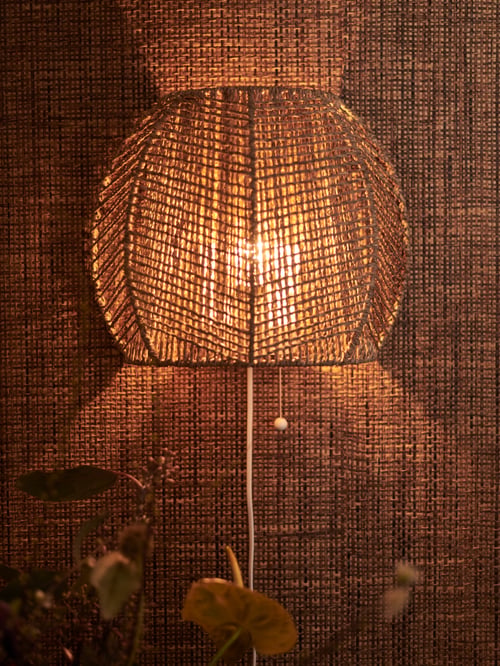 A MÅNALG wall lamp with flowers below it, on a wall with a natural covering. The bulb is on, creating a dramatic effect.