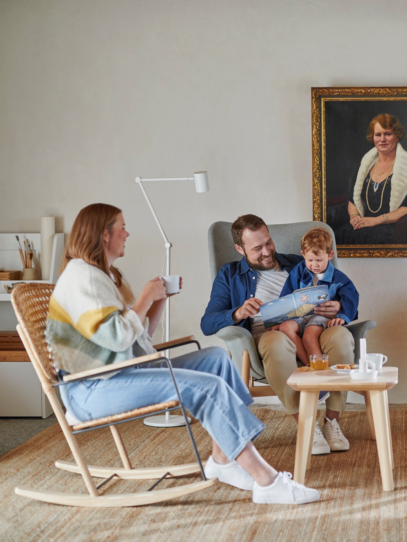 Two grown-ups and a child are lounging. The child is sitting on the lap, of a one of the grown-ups, reading a book.