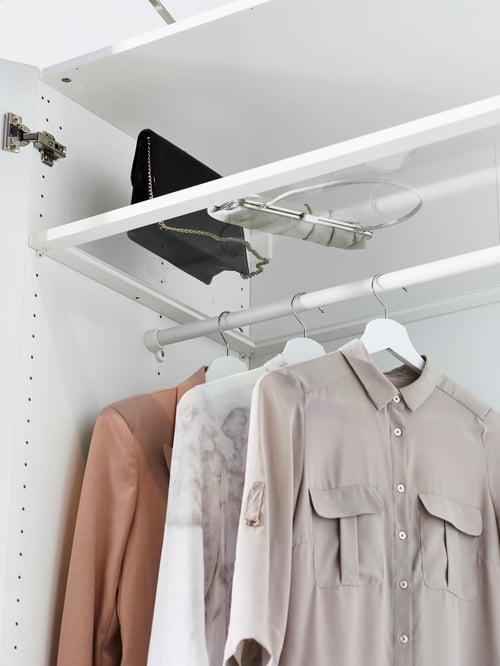 À l’intérieur d’une armoire-penderie, des vêtements sont suspendus à une tringle sous une tablette en verre KOMPLEMENT avec un cadre blanc, où sont rangés des sacs à main.