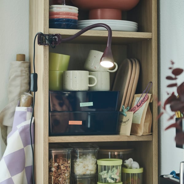 A NÄVLINGE LED spotligt is clamped onto a BILLY shelf containing ceramic mugs, trays and glass jars of dried foods.