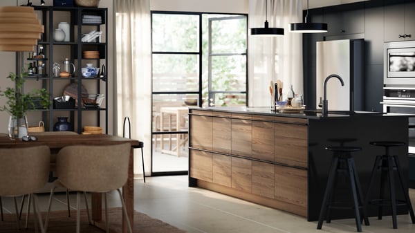 A bright kitchen with a STENSTA dark brown ash veneer drawer fronts, black bar stools, open shelving, and a dining table.