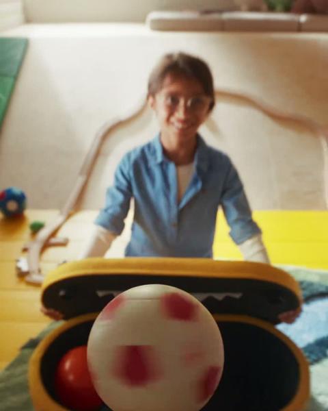 A mouth with an upper row of pointed teeth opens to reveal a young girl. The mouth opens and closes, and the girl throws toys into it.