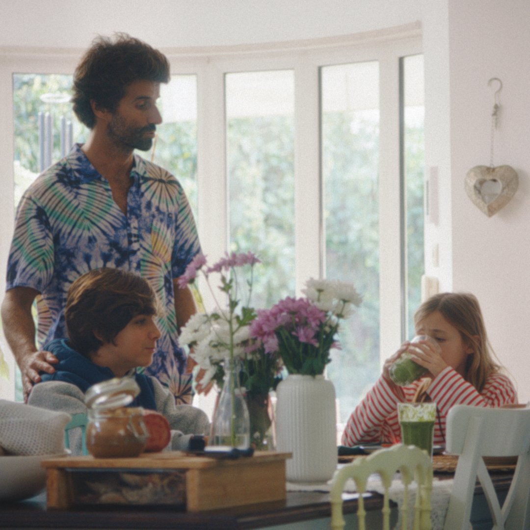 Eine erwachsene Person und drei Kinder sitzen in einem hellen Raum an einem Esstisch und trinken frisch zubereitete grüne Smoothies.