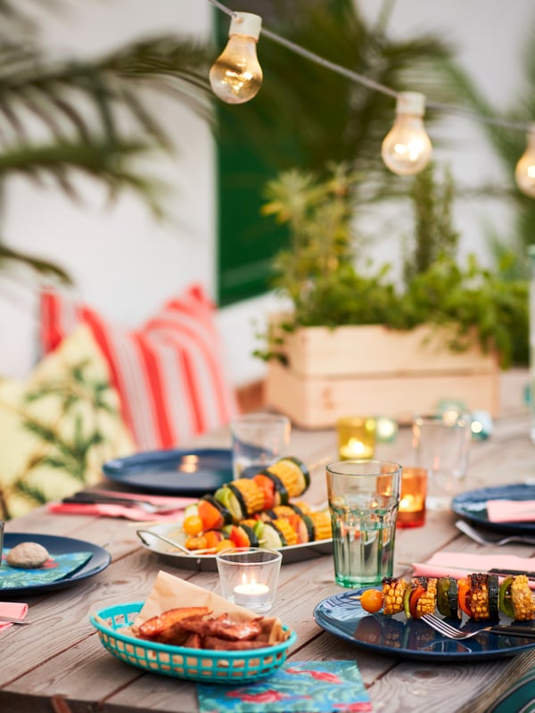 A wooden outdoor table surrounded by plants, lighting chains and textiles, set with FÄRGRIK plates and LIVNÄRA cutlery.