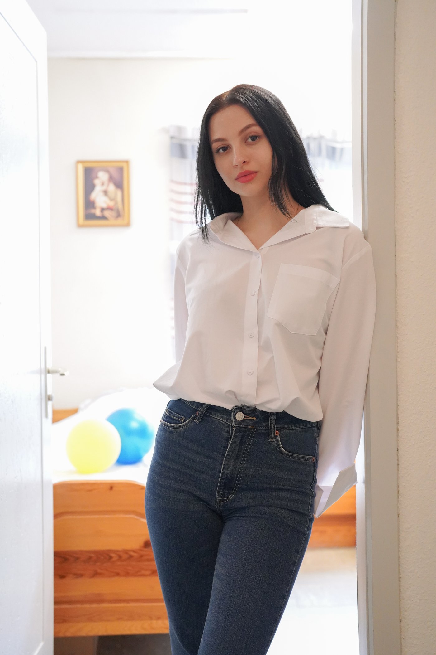 Girl leans against the doorframe in her living room and wears jeans and a white shirt.