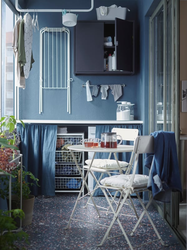 A balcony with a white table and two chairs, a foldable drying rack on the wall, and some plants.