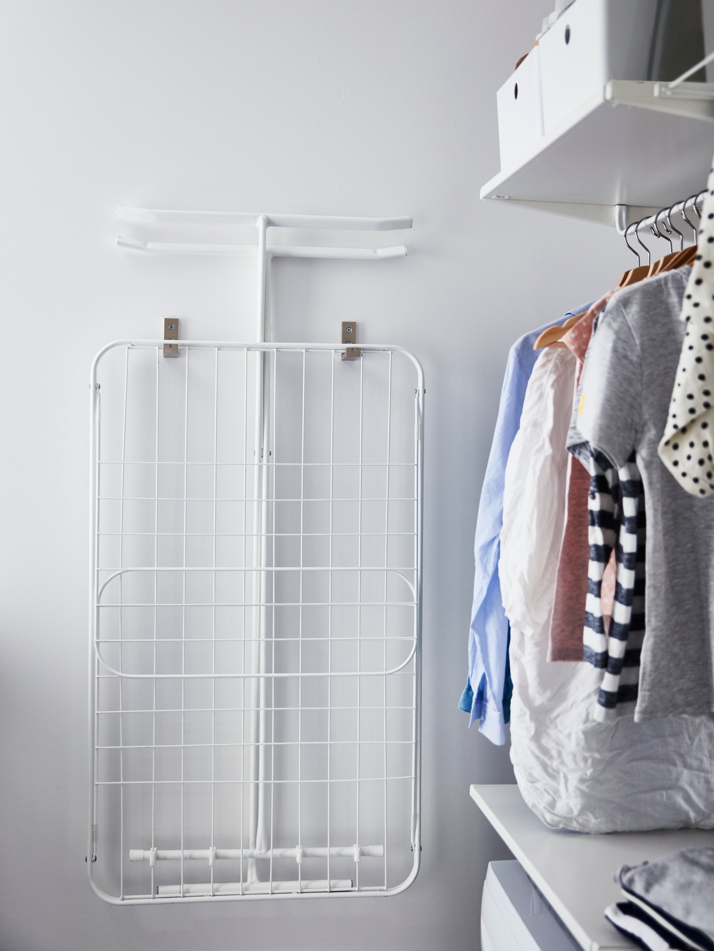 Un tendedero MULIG doblado y colgado en un muro blanco junto a ropa limpia y ordenada. 