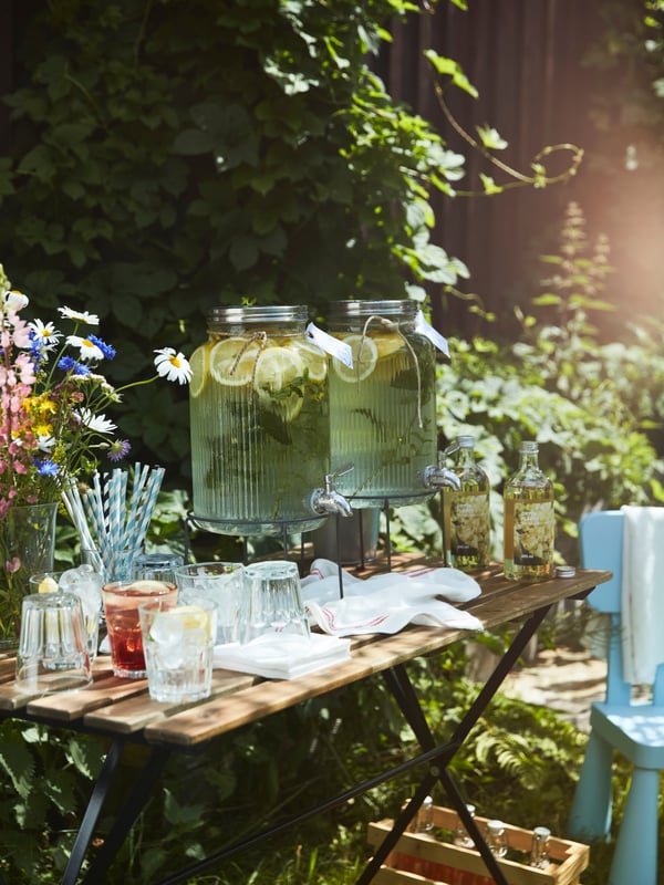 Ein kleiner Gartentisch mit Gläsern, Strohhalmen und Limonade in einem VARDAGEN-Glas mit Zapfhahn, bereit für die Gäste einer Party.