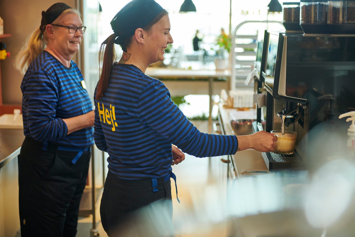 Kaksi sinisiin raitapaitoihin sonnustautunutta IKEA-työntekijää IKEA Ravintolassa. Toinen työntekijä ottaa juomaa kahviautomaatista.