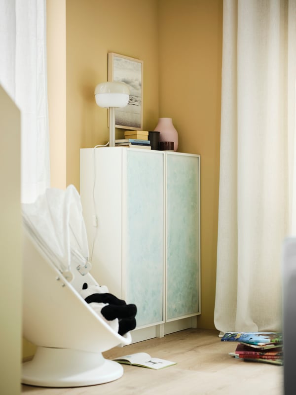 A BILLY/HÖGBO bookcase combination with glass doors with artwork on the panels is placed in the corner of a yellow room.