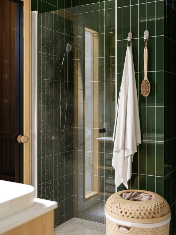 A shower is placed in a green-tiled bathroom and there is a white FREDIKSJÖN towel hanging from hooks on the wall next to it.