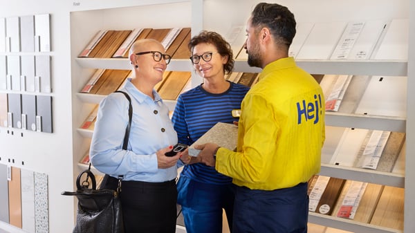 A smiling business customer in an IKEA store stands in front of shelves with material samples and is being consulted by an interior designer and her assistant.