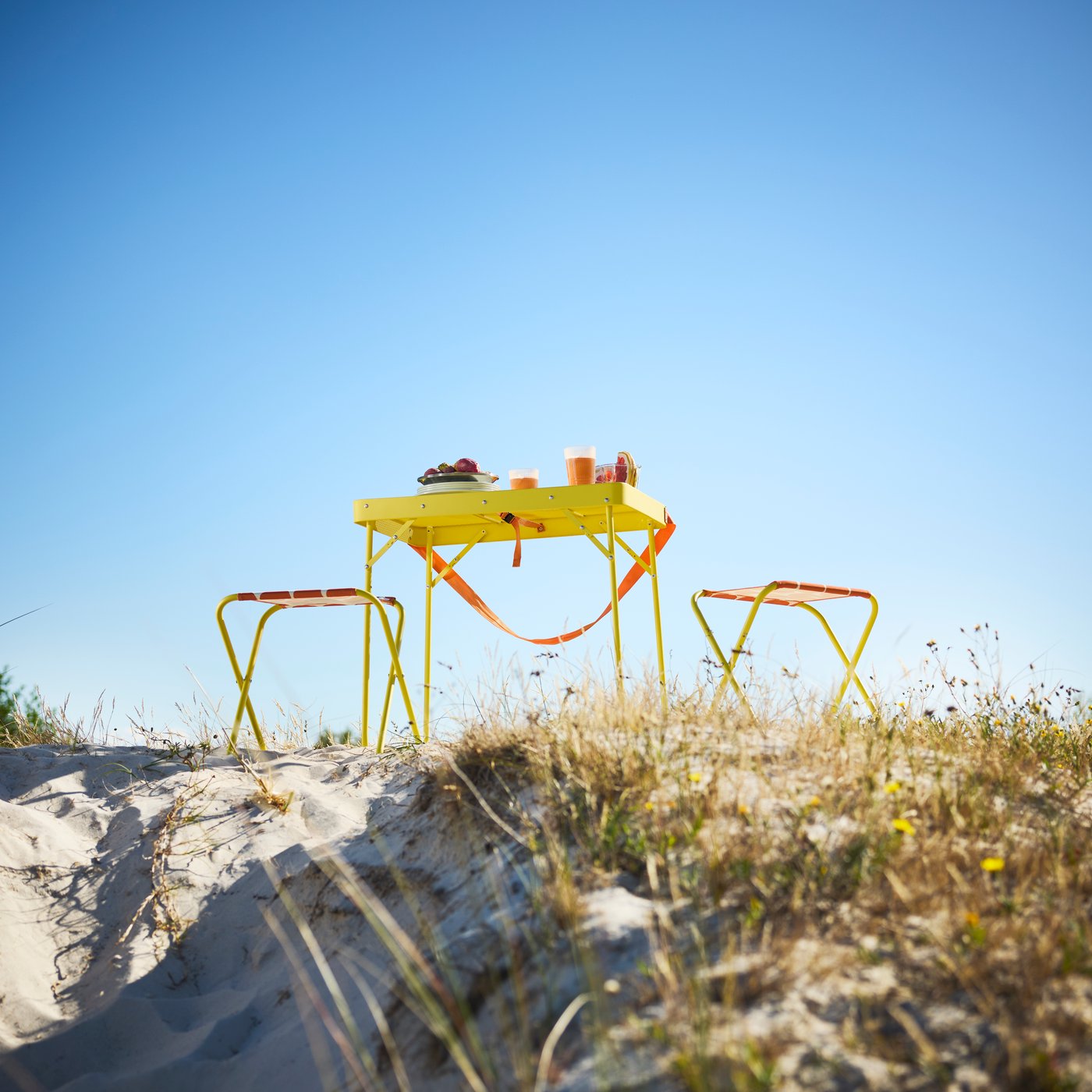 A beach scene features a bright yellow orange STRANDÖN folding table with two matching stools set on sand.