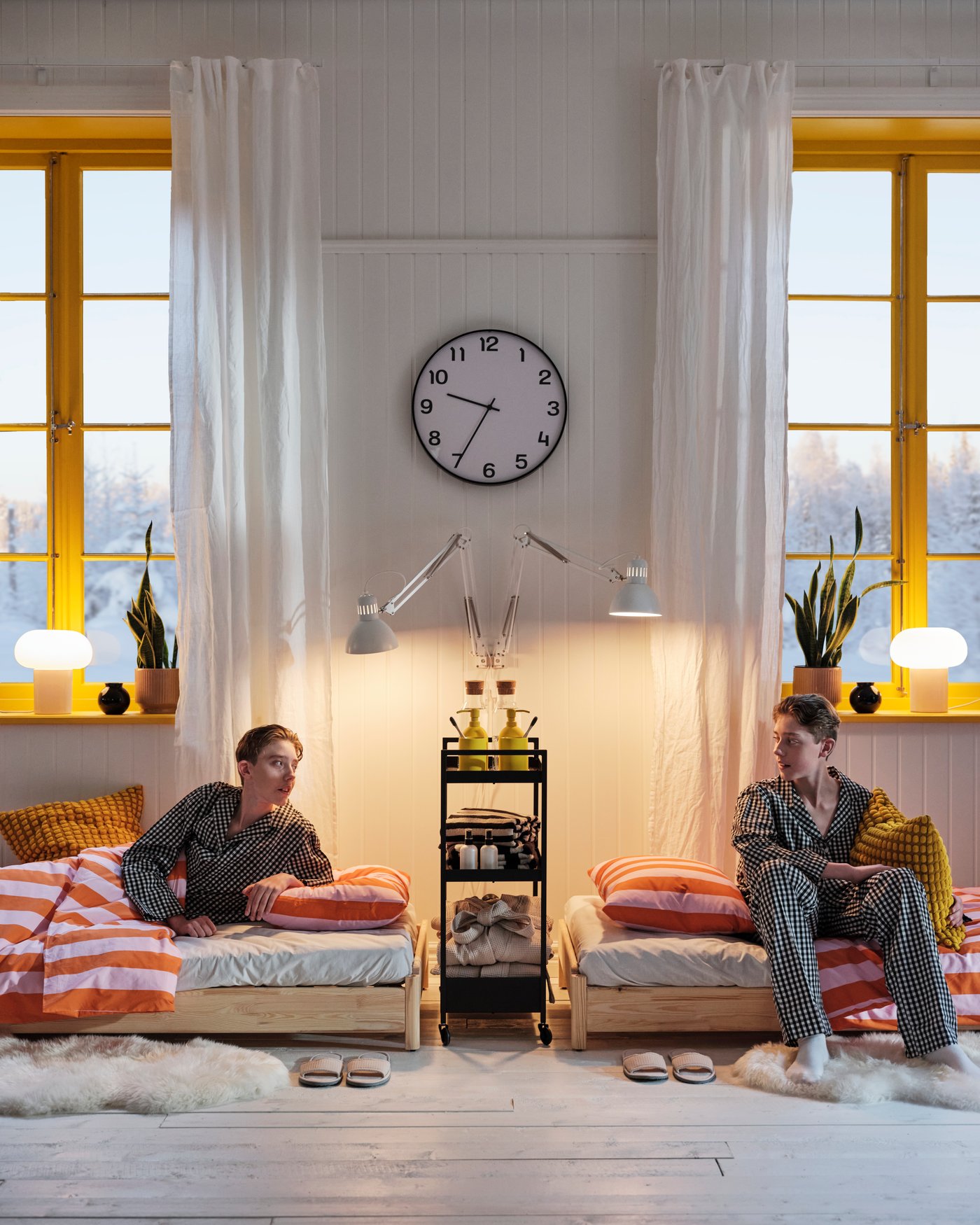 Two people sitting on UTÅKER beds divided by a trolley in a room with bright yellow windows.