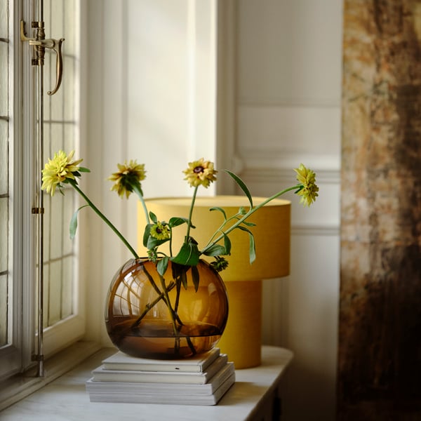 An amber-coloured STOCKHOLM 2025 vase and a yellow linen-covered STOCKHOLM 2025 table lamp
