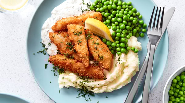 Five pieces of breaded fish bites, fried golden-brown, with mashed potatoes, green peas and a dip are placed on a blue plate.