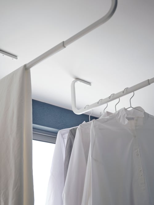 Laundry room with white wall, white clothes bars attached on ceiling, white shirts placed on white hangers, displayed on bar. 