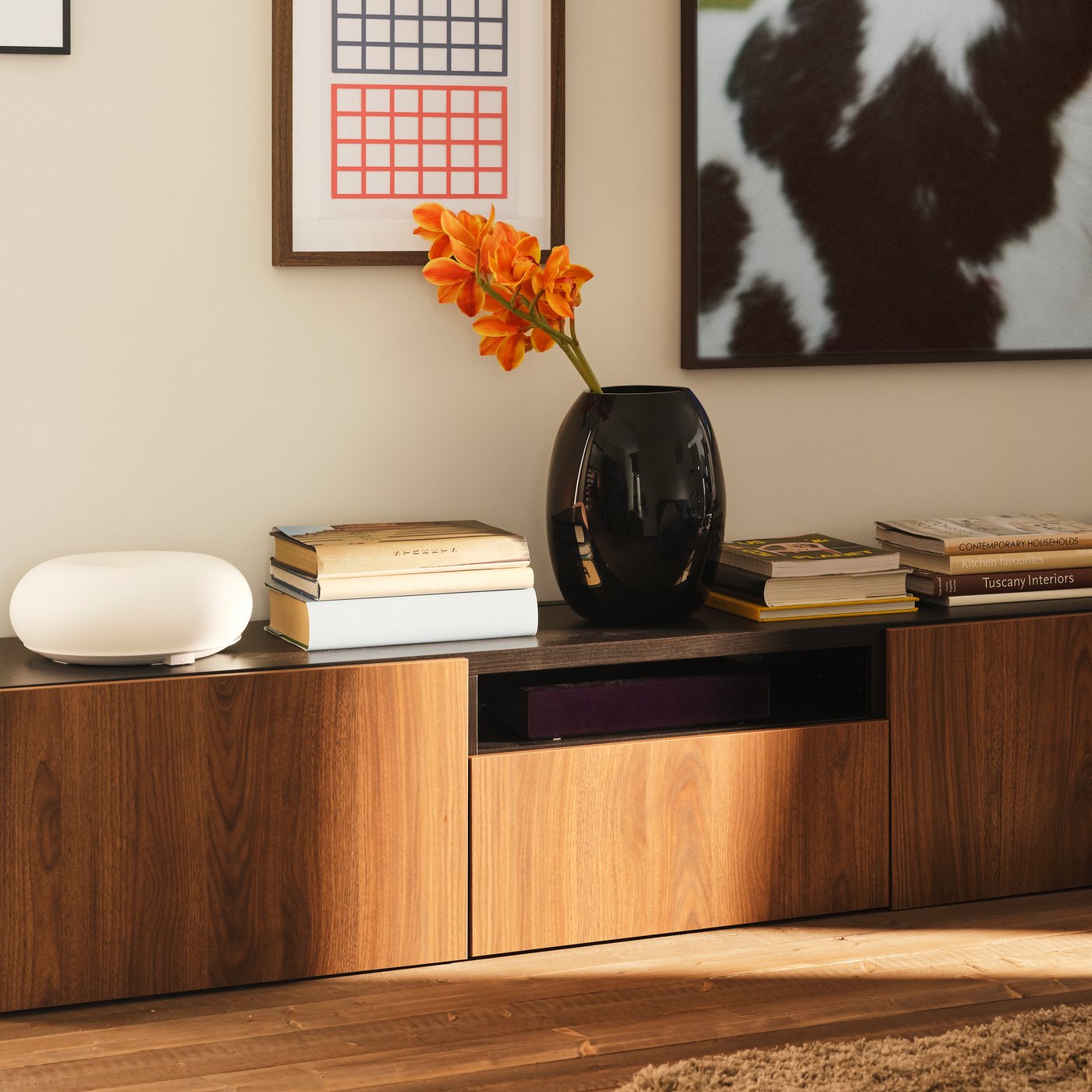 A lot of books and a black STOCKHOLM 2025 vase with flowers in it are on top of a BESTÅ TV bench standing against a wall.