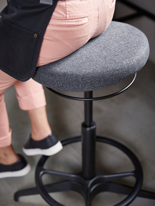 Persoană cu șorț în spațiul de lucru modern, așezată pe un suport de culoare gri închis, cu cadru metalic și pernă rotundă.