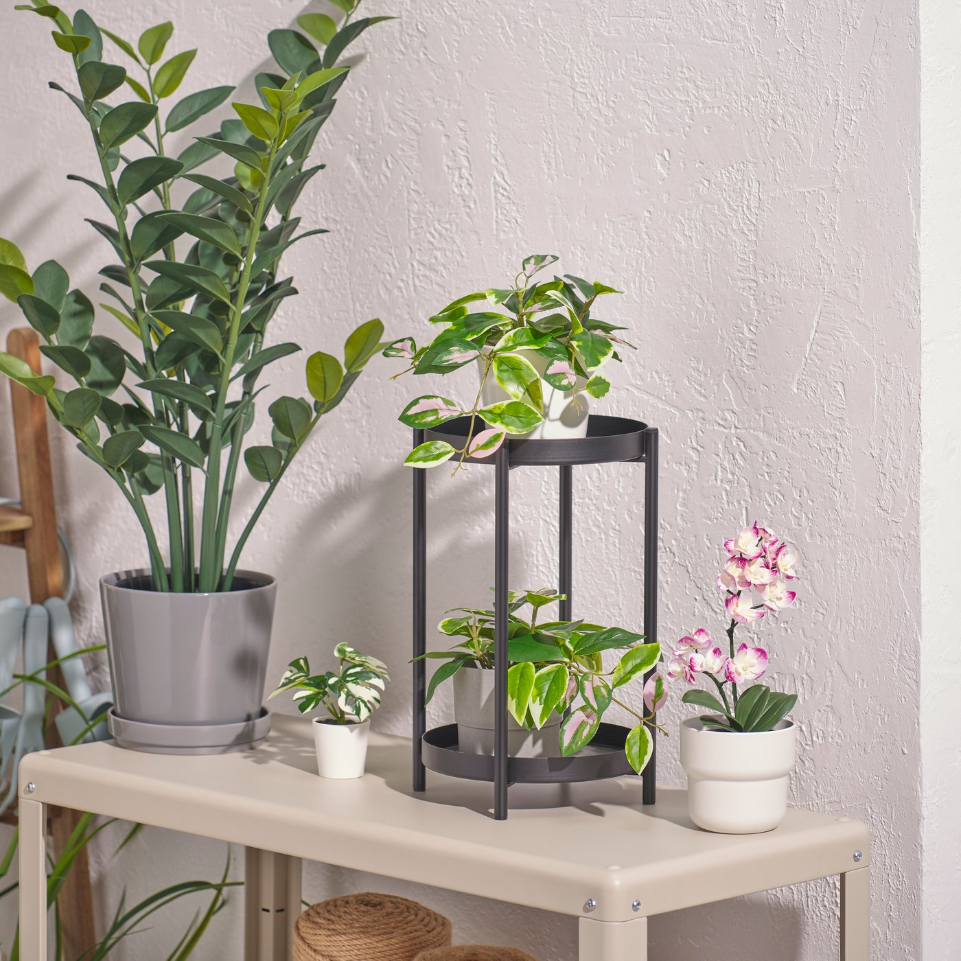 Piccola pianta artificiale FEJKA con fiori di orchidea lilla chiaro in vaso bianco, appoggiata sul ripiano inferiore di un carrello accanto ad altre piante artificiali in vaso.