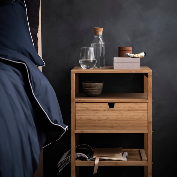A NORDKISA wooden nightstand with a glass and bottle of water. A bowl rests inside the nightstand with a square and circular container sitting on top of it.