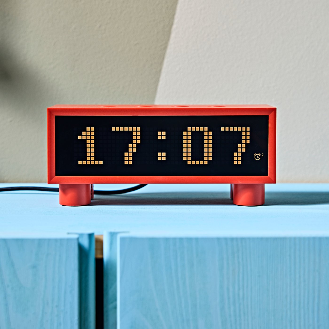 A close-up of a red, retro-style KORTLINJE alarm clock displaying 17:07, placed on a light blue cabinet by a beige wall.