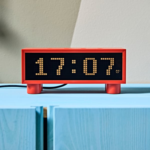 A close-up of a red, retro-style KORTLINJE alarm clock displaying 17:07, placed on a light blue cabinet by a beige wall.