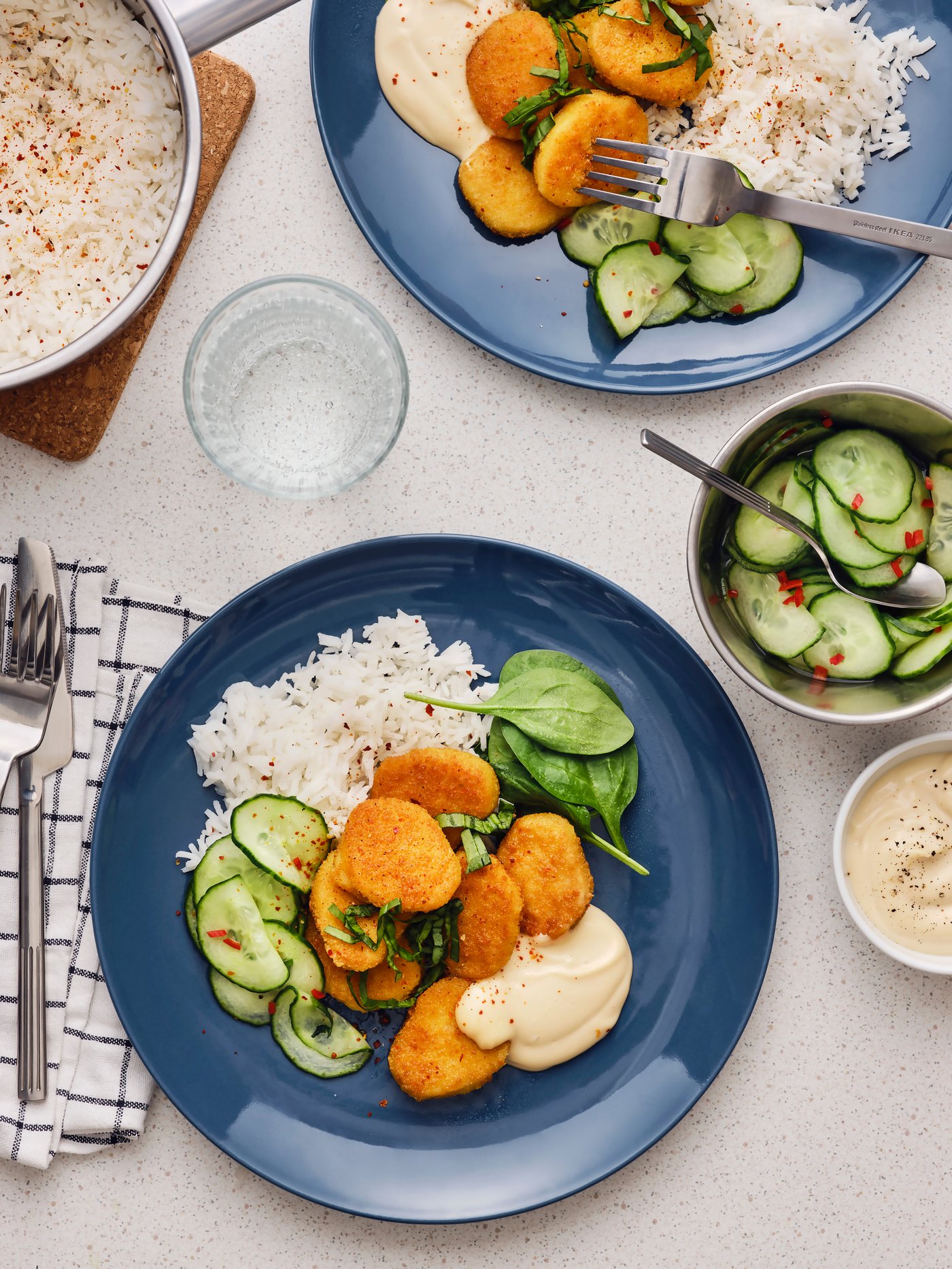 Auf einem Tisch mit Tellern ist ein Gericht aus Reis, Gurkensalat, panierten Häppchen und Spinatblättern zu sehen. Außerdem gibt es hier ein Glas Wasser und Besteck.
