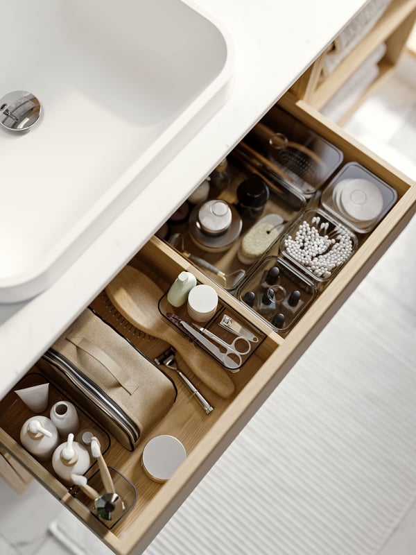 A bathroom vanity has a drawer that’s open and there are care products organized in grey VISSLAÅN boxes with lids inside.