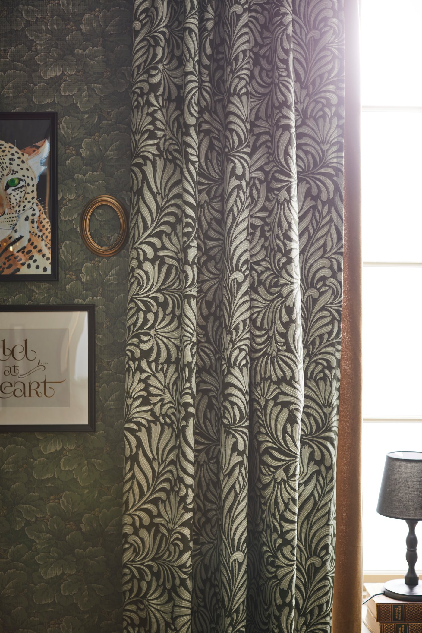 Tenda TRÅGSPINNARE verde scuro con fantasia a foglia in una stanza con cornici nere KNOPPÄNG alla parete e lampada ta tavolo