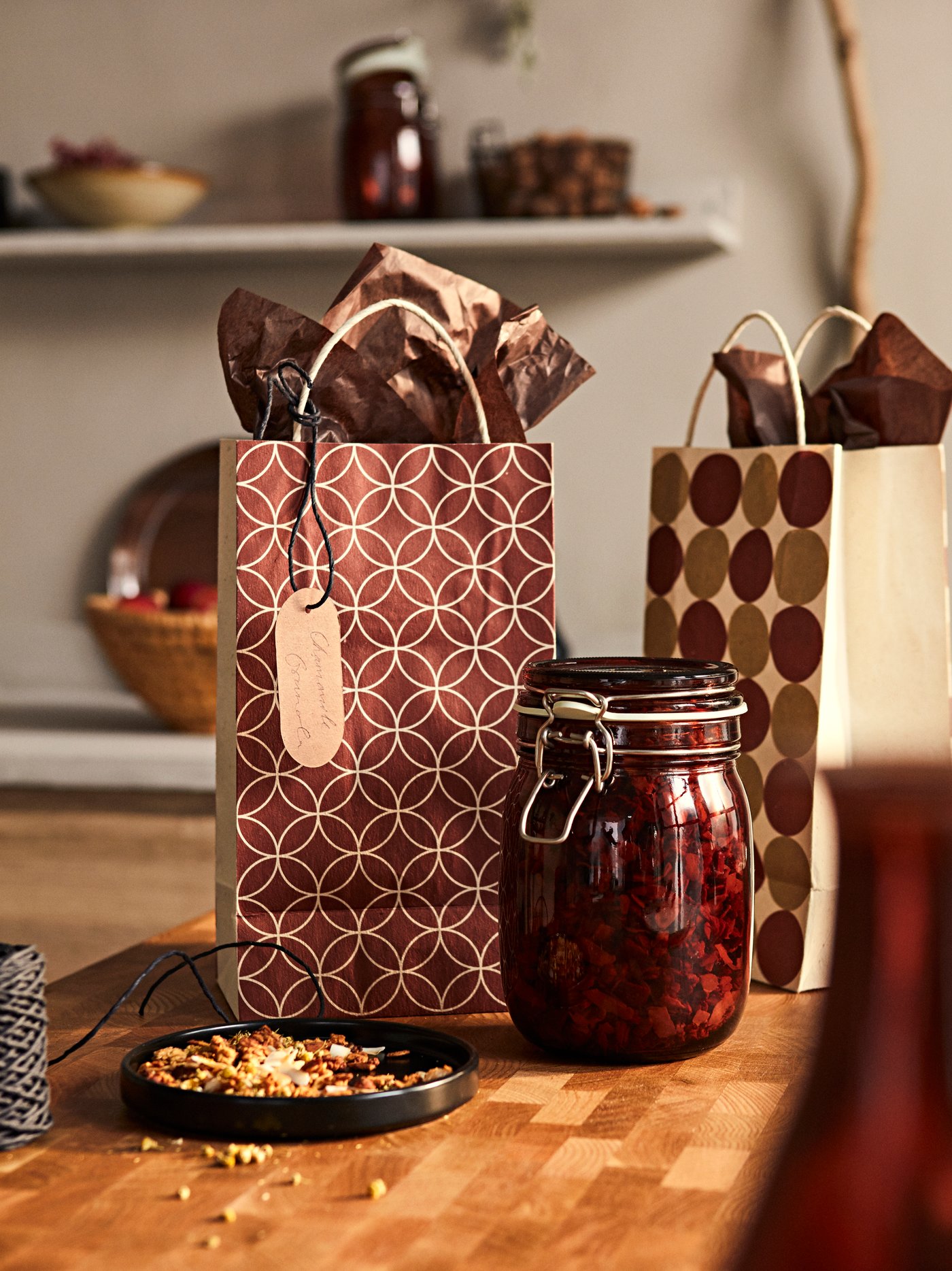 Dua beg hadiah KRÖSAMOS yang berlabel dan kertas pembalut di samping balang kaca berpenutup KRÖSAMOS yang berisi campuran teh herba.