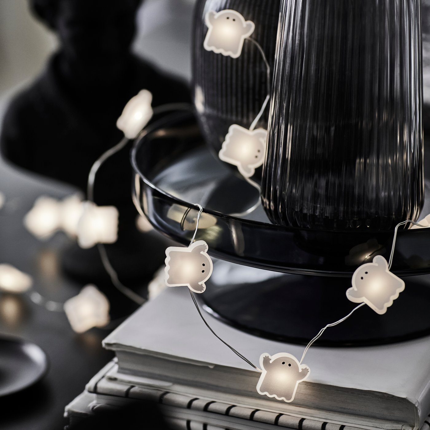 A KUSTFYR light chain featuring several small ghosts is wrapped around a black decoration and placed on top of some books.